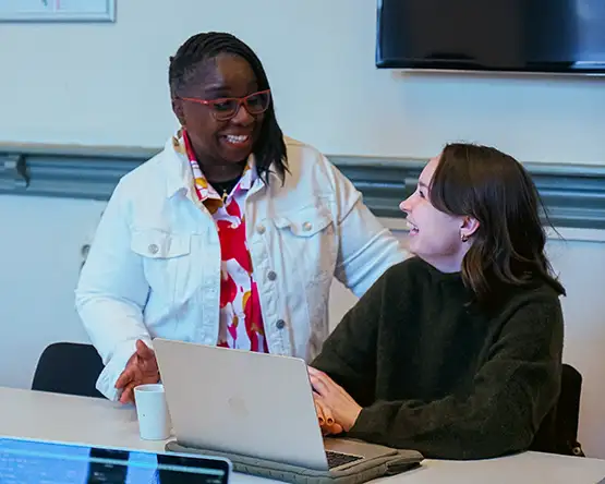 Student seated at a desk with laptop with a professor standing beside her.
